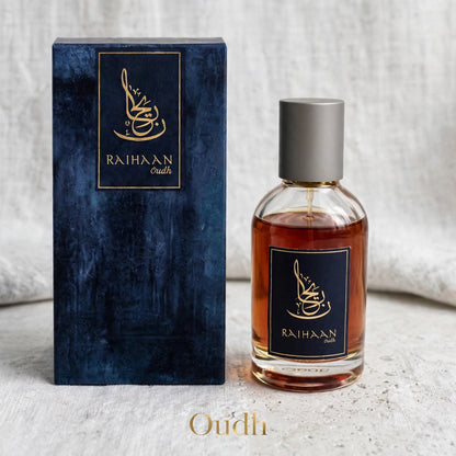 A bottle of Raihaan Oudh perfume with amber liquid and a silver cap stands next to its textured dark blue box with gold lettering, on a light fabric surface.