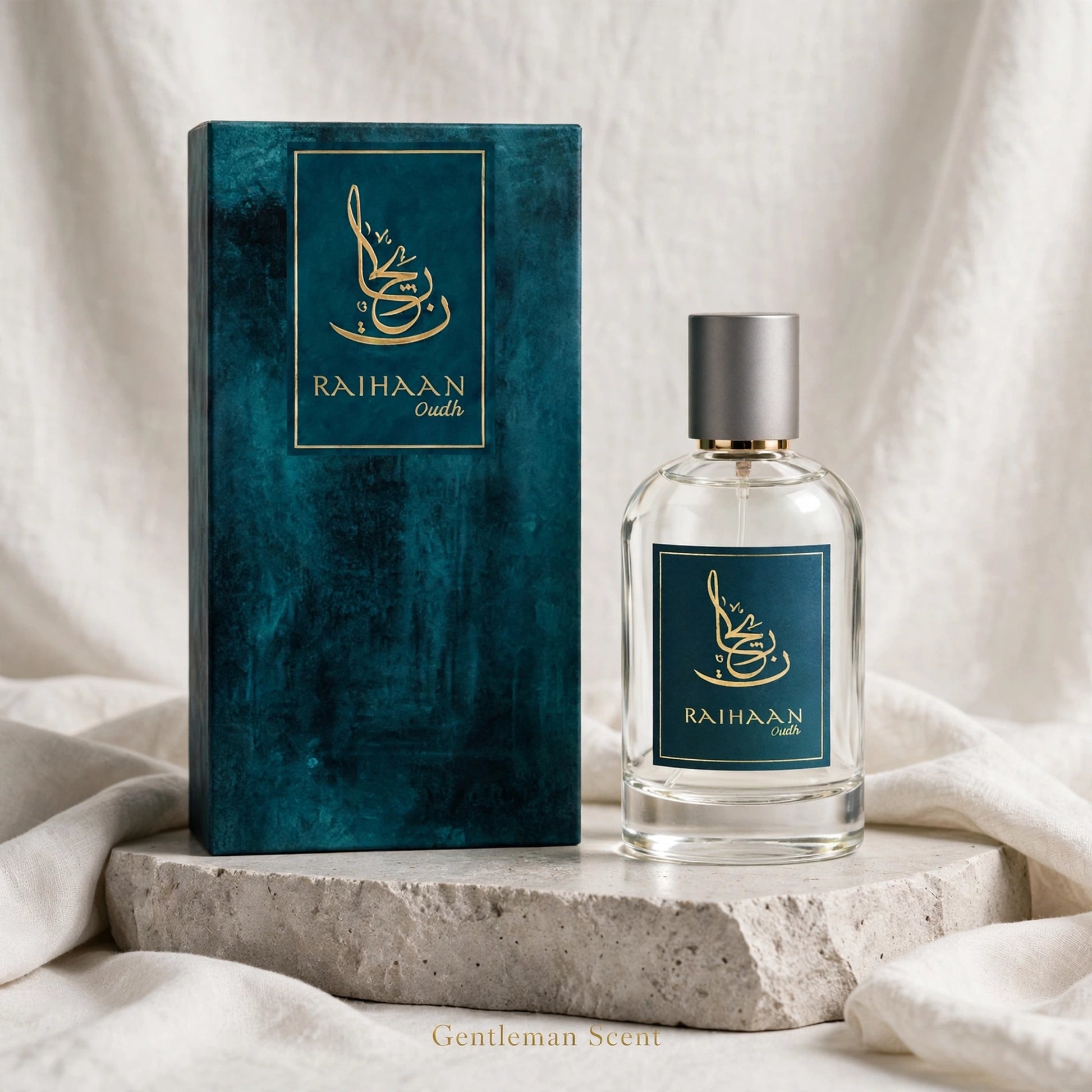 A clear glass perfume bottle with a silver cap stands beside a tall textured teal box with gold lettering reading RAHHAAN scents, set on a stone slab with draped white fabric in the background.