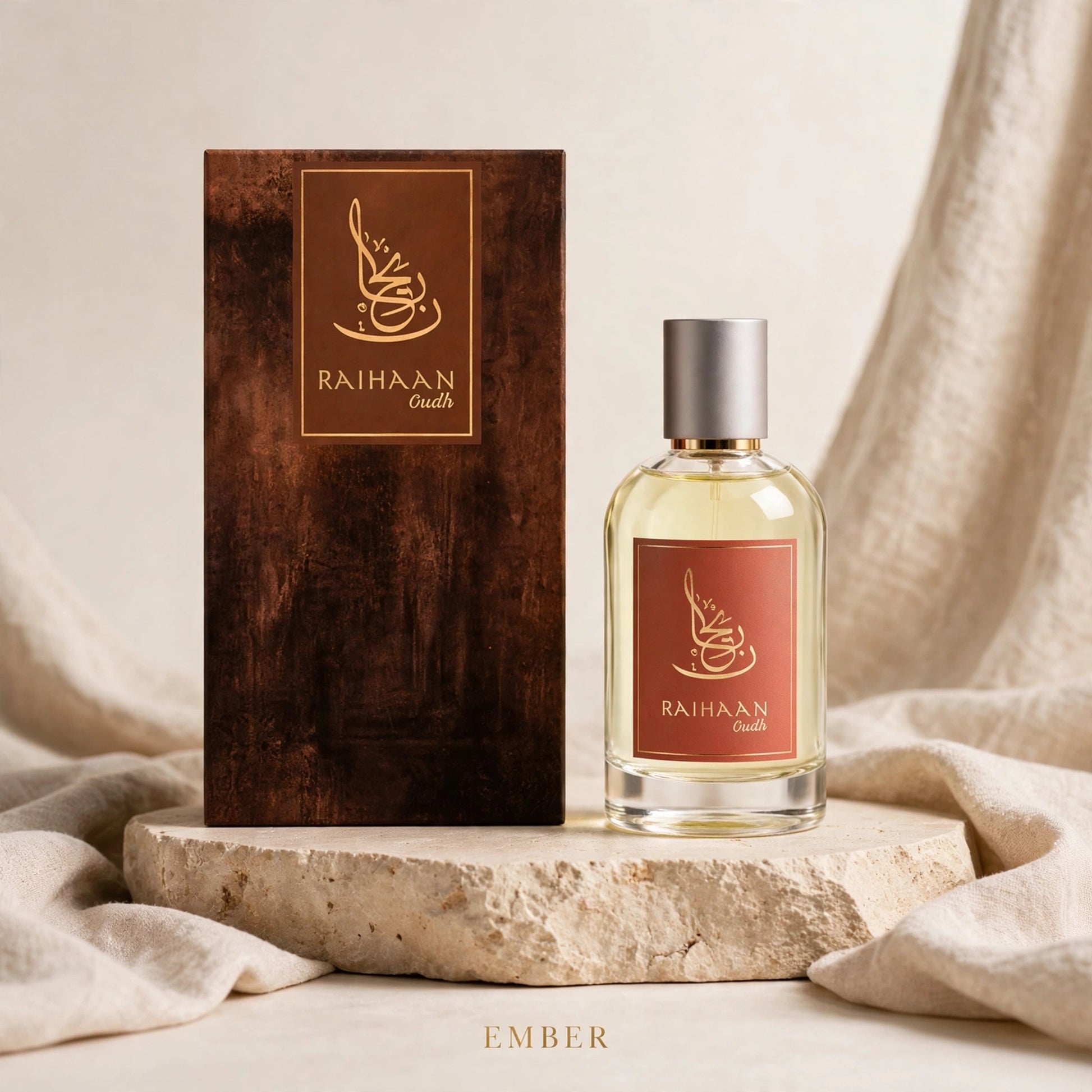 A bottle of Raihaan Gouti 'Ember' fragrance next to its textured dark brown wooden box, displayed on a stone slab with draped beige fabric.